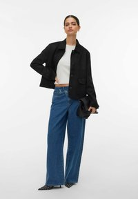 Veste courte noire, haut blanc, jean large bleu. Pochette noire, talons pointus à pois blancs. Tissu lisse, coupe décontractée.