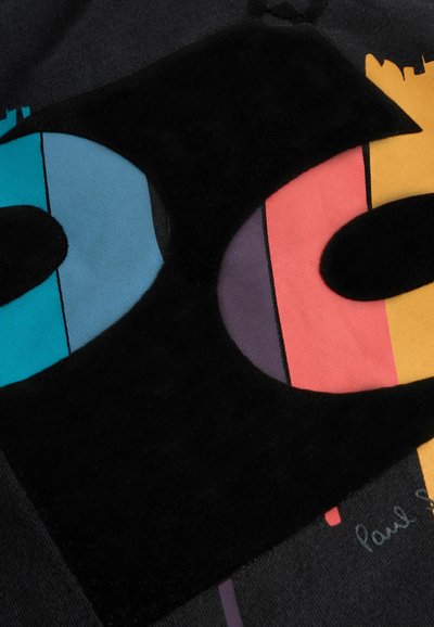 Mustat hupparit, joissa on suuri graafinen painatus, joka esittää tyyliteltyjä kirjaimia sinisessä, vaaleanpunaisessa ja oranssissa, painettuna teksturoituun kankaaseen. Näkyvissä on myös allekirjoitusyksityiskohta.