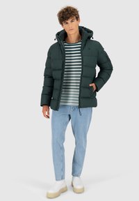 Giacca verde imbottita con cappuccio, maniche lunghe, zip frontale e tasche laterali. Indossata sopra una camicia a righe e abbinata a jeans azzurri chiari.