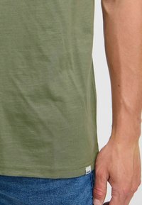 Camiseta verde oliva de tejido suave, con un corte recto y costuras visibles. Etiqueta con "SOLID" en el lado inferior, combinada con vaqueros azules.