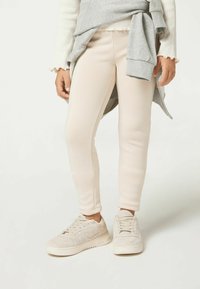 Beżowe legginsy w dopasowanym kroju o gładkiej fakturze, zestawione z jasnoszarymi trampkami o miękkiej, teksturowanej powierzchni.