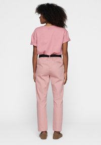 Camiseta de manga corta rosa claro con mangas enrolladas, combinada con pantalones rosa claro y una cinturilla negra. Modelo de espaldas, usando zapatillas suaves.