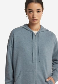 Jasnoniebieska bluza z zamkiem błyskawicznym i kapturem ze sznurkiem, z ryflowanymi detalami oraz kieszeniami z przodu; wykonana z miękkiego materiału o teksturowanym wykończeniu.