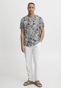 Cămașă cu mâneci scurte, cu nasturi, având un model floral albastru și bej, confecționată dintr-un material ușor, asortată cu pantaloni drepti albi și sandale maro.