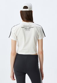 Camiseta blanca cortada con acentos negros, que presenta texto en la parte posterior y mangas cortas. Combinada con leggings negros ajustados.