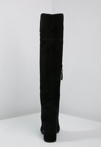 Bota de ante negra hasta la rodilla con tacón de bloque y cremallera lateral, mostrada desde la parte posterior sobre un fondo gris liso.