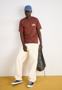 T-shirt en coton marron avec le logo "Dickies", pantalon large crème, baskets navy et casquette bleu clair. Tient un sac texturé foncé.
