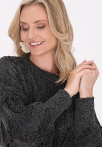 Pulover texturat, de culoare gri închis, cu o croială lejeră și mâneci lungi. Prim-plan cu mâini ușor împreunate, evidențiind materialul moale din tricot.