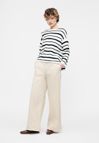 Άτομο που φοράει μακρυμάνικη μπλούζα με ασπρόμαυρες ρίγες, μπεζ παντελόνι με φαρδιά μπατζάκια και σκούρα καφέ παπούτσια, στέκεται με τα χέρια στις τσέπες.