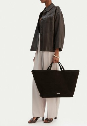 Frau trägt dunkelbraune Lederjacke und beige Hosen mit weitem Bein, hält eine große schwarze Tote-Bag mit zwei Griffen und dezentem Logo.