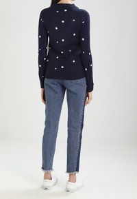 Granatowy sweter z białymi geometrycznymi wzorami, zestawiony z jasnoniebieskimi dżinsami z postrzępionym obszyciem oraz białymi trampkami. Klasyczny codzienny strój.