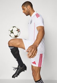 Hvid fodboldtrøje med røde detaljer, kombineret med sorte shorts og knæhøje sorte sokker. Spilleren balancerer en fodbold på sit knæ.