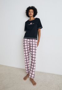 T-shirt preta de algodão com texto bordado e calças xadrez cor-de-rosa com linhas de grelha pretas. Conjunto exibido sobre um fundo de carpete claro.