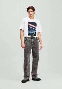 Jovem vestindo uma t-shirt branca com estampa gráfica, jeans retos cinza desbotado e sapatos pretos, em pé contra um fundo simples.