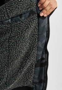 Fleece foret vest med en struktureret grå ydre lag, der har et camouflageprint indvendigt, lynlåslukning og sidelomme.
