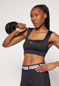 Sujetador deportivo negro con escote cuadrado, que presenta un logo en el frontal y una banda con marca. Material suave, combinado con leggings de cintura alta.