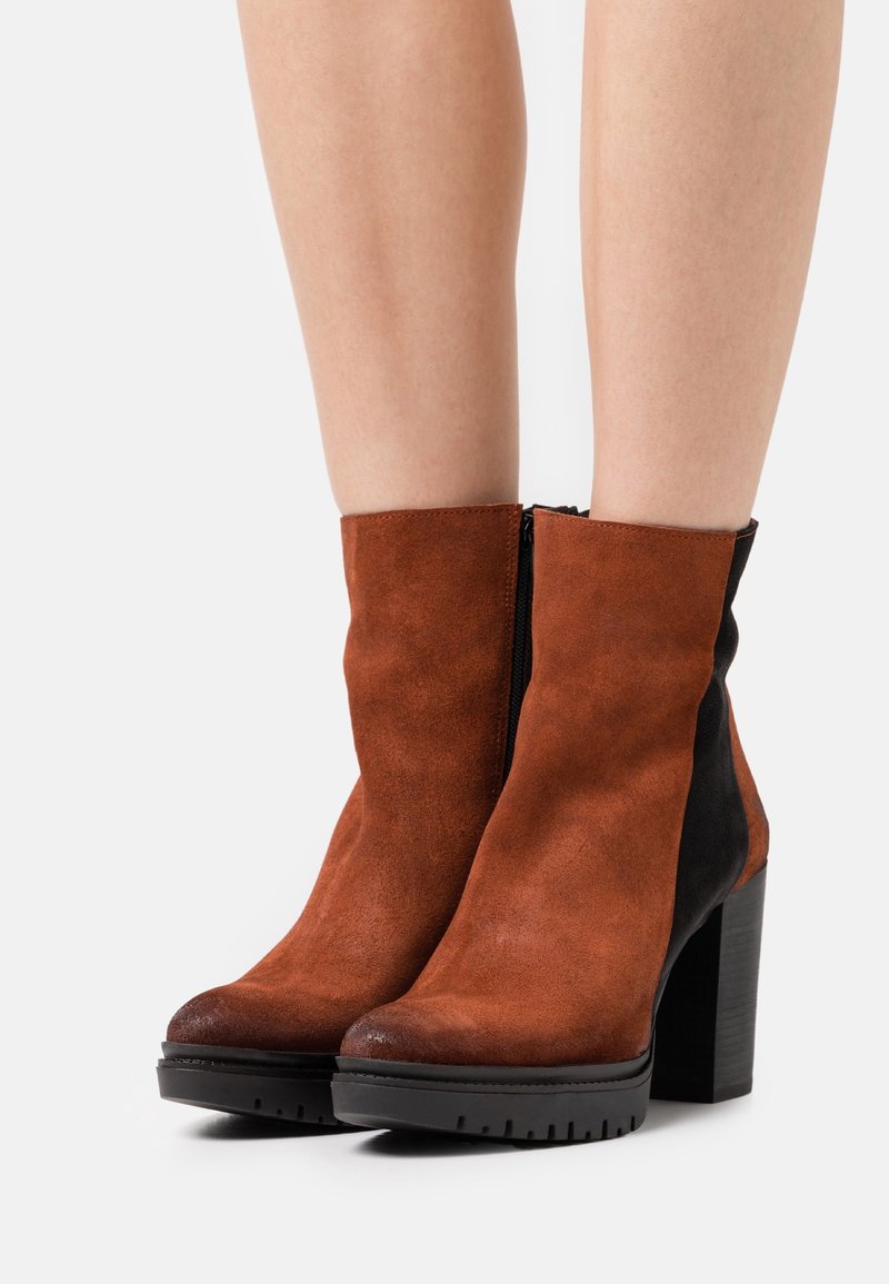 Bottes en daim montant à la cheville avec l'avant marron et l'arrière noir, talons blocs épais, portées sur des jambes nues contre un fond uni.