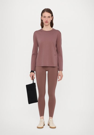 Langärmliges mauve Oberteil mit runder Halslinie, kombiniert mit passenden Leggings. Schwarze Clutch in der Hand, cremefarbene Sneakers setzen einen Kontrast zum Outfit.