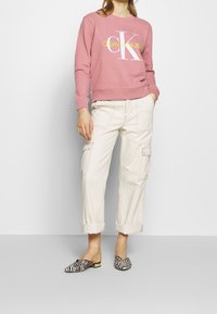 Sudadera rosa con logotipo blanco y amarillo, pantalones cargo color crema con puños arremangados y zapatos de lona de patrón blanco y negro.
