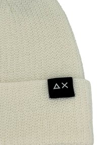Beanie lavorato a maglia color crema con risvolto; presenta un'etichetta nera con logo geometrico bianco sul davanti. Materiale testurizzato.
