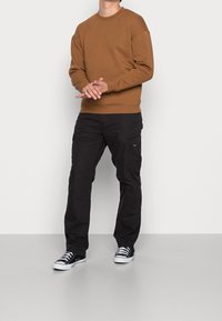 Felpa marrone con scollatura rotonda, abbinata a pantaloni cargo neri con tasche laterali e sneakers nere con dettagli bianchi.
