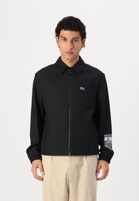 Black zip-up jacket with a collar, textured fabric, featuring a chest pocket and a graphic accent on the sleeve. Elastic cuffs.
