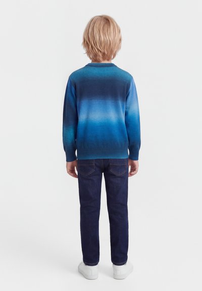 Ξανθό παιδί όρθιο, γυρισμένο με την πλάτη, φοράει μπλε πουλόβερ με διαβάθμιση, σκουρόχρωμα τζιν και λευκά αθλητικά παπούτσια σε απλό φόντο.