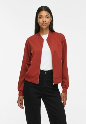 Donna con capelli lunghi e scuri che indossa una giacca con zip color ruggine, camicia bianca e pantaloni neri a vita alta su uno sfondo chiaro uniforme.