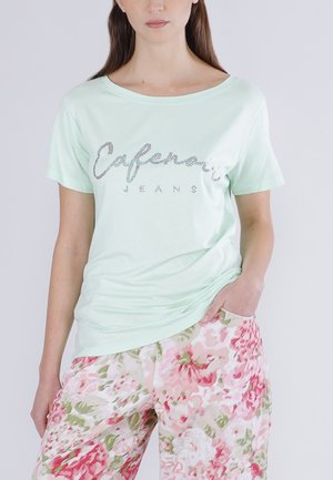 Kvinde iført mintgrøn "Cafenoir Jeans" t-shirt og løstsiddende blomstrede bukser med pink, hvide og grønne blomster, med den ene hånd i lommen.