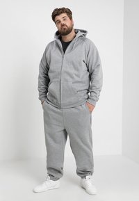 Sudadera con cremallera de color gris claro y pantalones de chándal a juego, confeccionados en un tejido suave, con bolsillos y puños elásticos, combinados con zapatillas blancas.