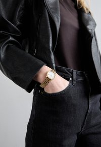 Goldene Uhr mit einem strukturierten Armband, weißem Ziffernblatt und römischen Ziffern. Schwarze Lederjacke über einem dunklen Hemd, kombiniert mit schwarzen Jeans.