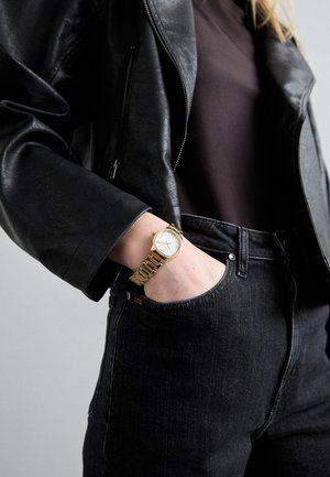 Orologio d'oro con cinturino testurizzato, quadrante bianco e numeri romani. Giacca di pelle nera sopra una maglietta scura, abbinata a jeans neri.