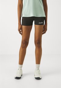 Pantaloni sportivi neri realizzati in un tessuto elasticizzato con una vestibilità aderente. Presentano un logo bianco sulla gamba sinistra. Indossati con una maglietta verde chiaro e scarpe bianche.