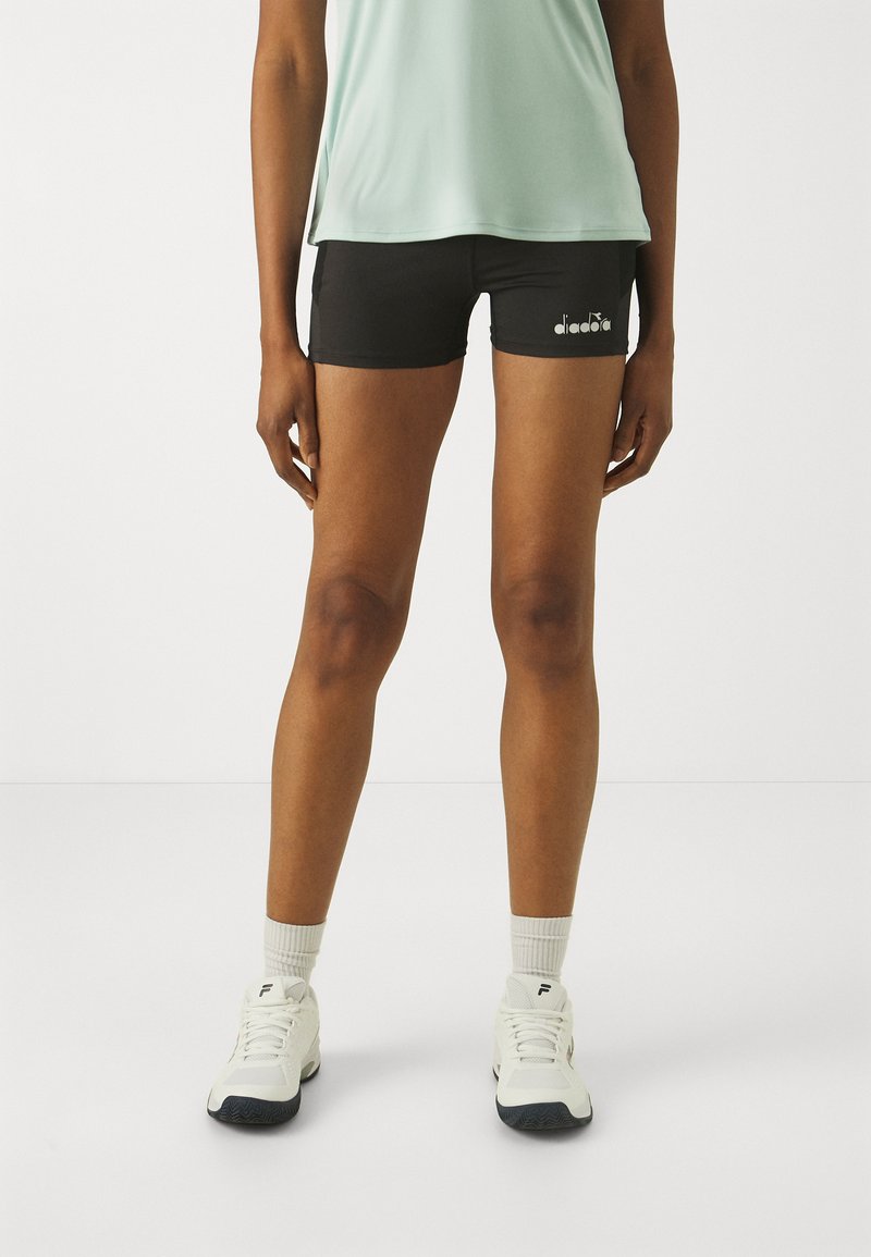 Pantaloni sportivi neri realizzati in un tessuto elasticizzato con una vestibilità aderente. Presentano un logo bianco sulla gamba sinistra. Indossati con una maglietta verde chiaro e scarpe bianche.