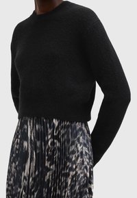 Personne portant un pull noir duveteux associé à une jupe plissée noire et blanche à motif abstrait, debout devant un fond uni.
