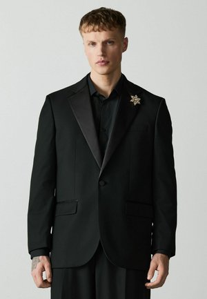 Young man with short hair wearing a black suit jacket and shirt with a gold star-shaped brooch on the left lapel against a plain background.