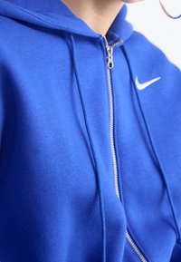 Sudadera con capucha azul con cremallera, hecha de tela suave con una textura lisa, que presenta un logo blanco de Nike en el pecho y una capucha ajustable.