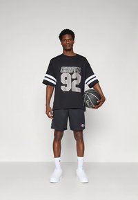 Svart oversized t-shirt med grå tryck "CHAMPION 92" och vita ränder på ärmarna, kombinerad med svarta shorts och vita sneakers, håller en basketboll.