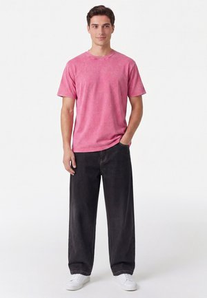 Giovane uomo in piedi con una mano in tasca, indossa una maglietta rosa a maniche corte scolorita, jeans neri larghi e sneakers bianche su sfondo bianco.