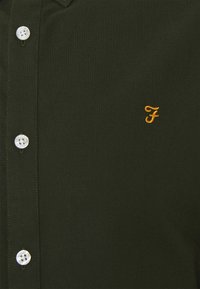 Chemise boutonnée vert foncé avec des boutons blancs et une petite lettre F jaune brodée sur la poitrine.