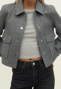 Szary żakiet w jodełkę wykonany z teksturowanej tkaniny, z kołnierzem, srebrnymi napami i dwoma przednimi kieszeniami. Uzupełniony szarym topem i ciemnymi dżinsami.