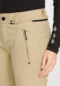 Beżowe spodnie narciarskie z teksturowanym wykończeniem, z suwakiem kieszeni przesuwnej, regulowanym pasem i haftowanym logo "Volcom".