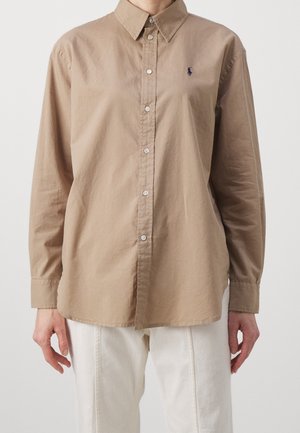 Person iført en beige langærmet knap-op-skjorte med et lille broderet logo på brystet og hvide bukser mod en ensfarvet baggrund.