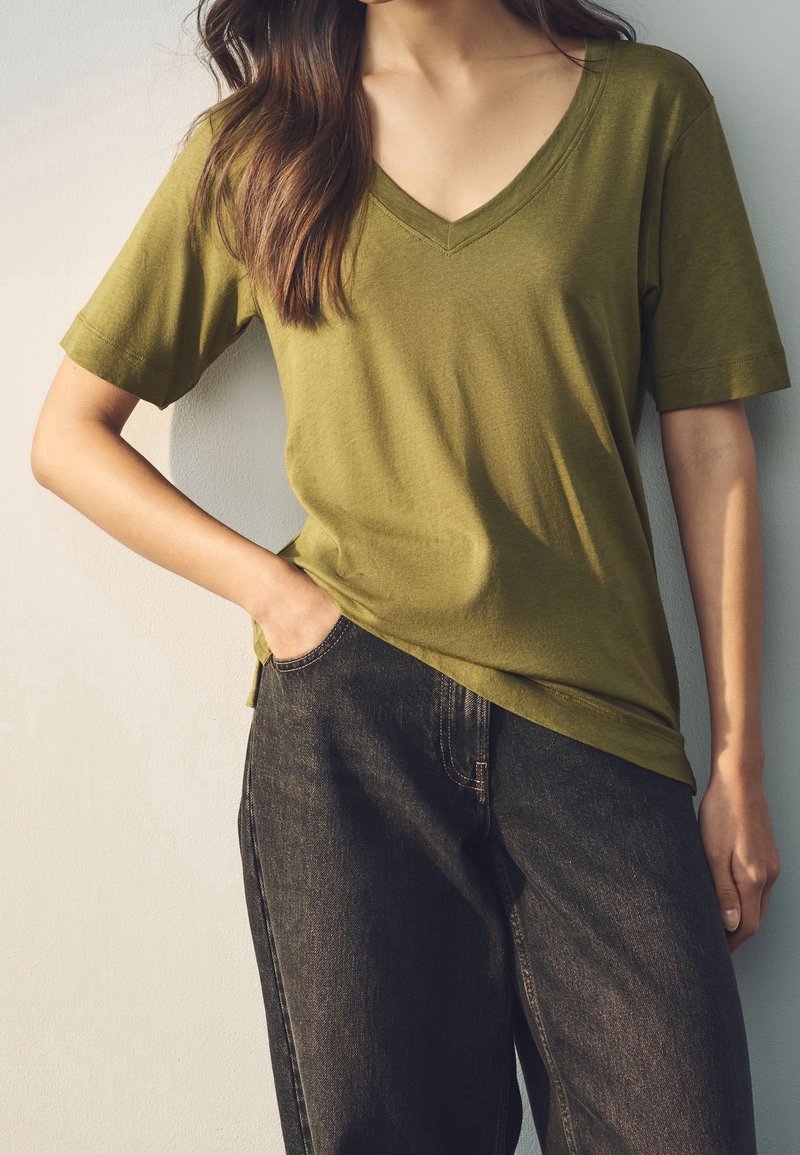 Femme portant un t-shirt vert olive à manches courtes avec col en V et un jean sombre ample, une main dans la poche, sur un fond clair uni.
