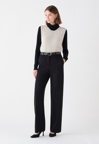 Gilet en tricot crème superposé sur un col roulé noir, associé à un pantalon noir taille haute et des escarpins noirs à bout pointu.