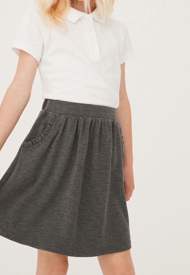 Enfant portant un polo blanc à manches courtes et une jupe gris foncé avec une taille froncée et des détails de poches à volants.