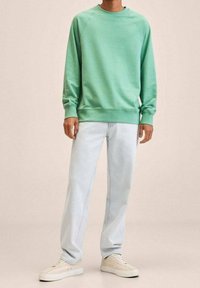 Felpa verde chiaro con polsini e orlo a costine, abbinata a jeans azzurri. Calzature sono scarpe da ginnastica beige dal design minimalista.