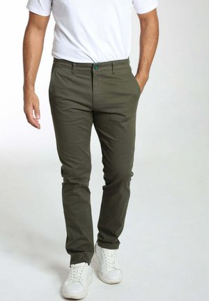 Homme portant un pantalon vert olive, des baskets blanches et un t-shirt blanc, debout avec une main dans la poche, sur un fond uni.