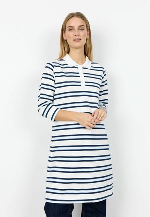 Kvinde med langt blondt hår iført en hvid kjole med marineblå og lyseblå vandrette striber, stående med hænderne foldet.