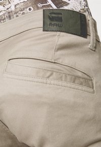 Poche de pantalon beige avec un patch en cuir noir étiqueté "RAW" près de la ceinture, et une partie d'une chemise à motifs rentrée.